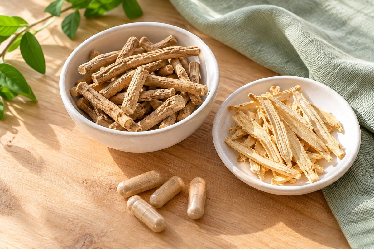 A white bowl of dried ashwagandha root pieces and a white dish of pale shatavari root slices with a few plain capsules on a light wood table